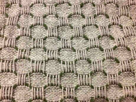 Handwoven cloth with a checkerboard pattern of contrasting textures in gray wool. One set of boxes are plain weave and the other are much looser vertical over weave. Between each row is a wavey contrasting line of green yarn, giving the whole thing a honeycomb effect.