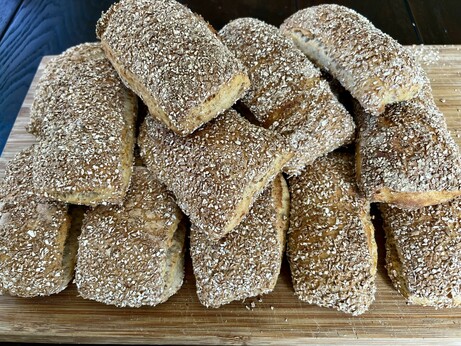 Auf einem dunklen Holztisch liegt ein Holzbrett. Darauf liegen zahlreiche frisch gebackene Brötchen mit rustikalem Ausbund.