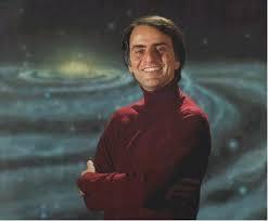 An image of Dr. Carl Sagan, wearing a red sweater, standing with his arms folded, in front of a luminous painted image of a great spiral galaxy.