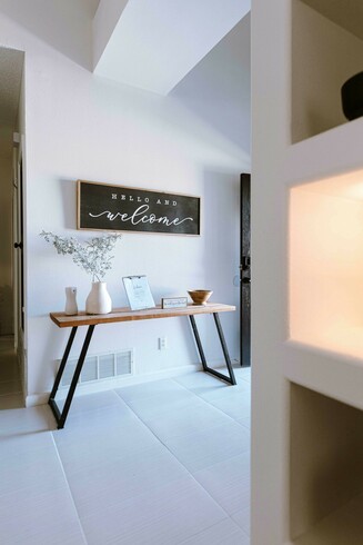 A photo of a modern-looking house, with a end table with a vase and other knick-knacks on it, with a hanging sign above it saying "Hello and Welcome". Photo by Rebeca Alvidrez on Unsplash