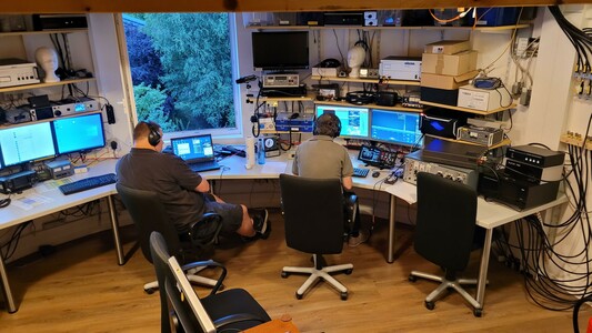 Amateurfunk Shack mit mehreren Computern und Funkgeräten. Am FlexRadio ein Operator und ein alter Contesthase im Stuhl daneben, der den Anfängern gute Tips gibt. #Trainer