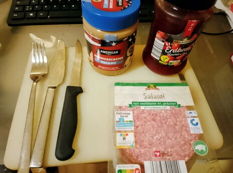 Creamy peanut butter, strawberry preserves, and salami wait on a white acrylic cutting board, along with cutlery.