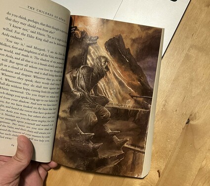 A person's hand holding open the book The Children of Hurin with the book open to an illustration of Hurin transfixed on a seat. The illustration looks scary.