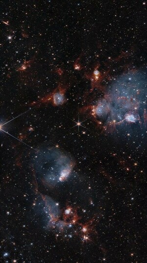 Bluish clouds of gas are wound through with red-glowing lanes and filaments of dust.. The clouds contain cavities filled with stars and many more stars are visible in the background and scattered throughout the clouds. The clouds are concentrated in the upper right and the lower middle of the image. Tiny background galaxies are also sprinkled throughout the image.