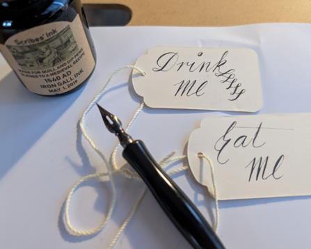 A pointed pen (hunt extra fine 512 in standard speedball nib holder) sitting in a mess of gift tag string with gift tags that say "Drink me" and "Eat me" in a passable, Spencerian-adjacent cursive. A bottle of ink sits near the top left of the image labeled "Scribes' Ink 1540 Iron Gall Ink"