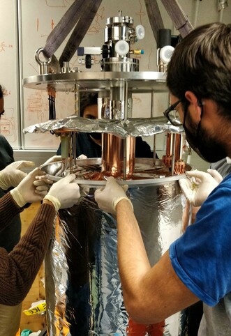 Three individuals wearing gloves assembling  a large metallic scientific apparatus with various components, in a laboratory setting.