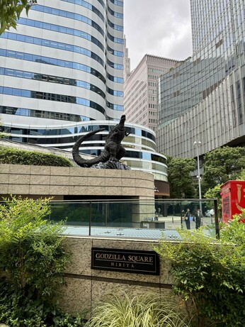 In a cluster of office buildings in downtown Tokyo is centered a wall, with a black bronze statue of Godzilla on top of it. Godzilla (the Shin Godzilla version) is shown roaring at the sky, arms and tail raised. In the foreground is a plaque that reads Godzilla Square - Hibiya in English.