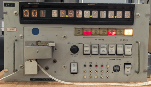 A front view of the Up-Data Link Test Set. It is a large rack-mountable box. It has a 13-digit display at the top. At the left is a paper-tape reader with a paper tape in it. To the right are various lights and switches.