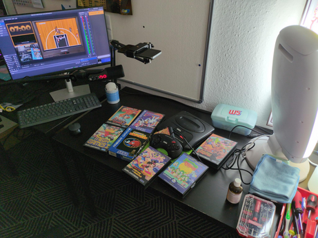 Photo of my capture setup. There is a table with the Mega Drive loot, tools and cleaning material on it. In addition there is a mobile phone as a camera, a microphone and a monitor showing OBS and the Owncast chat.