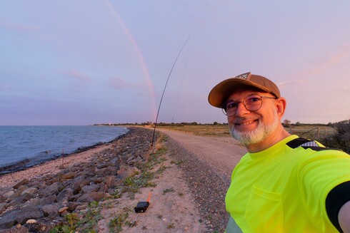 Selfie of DJ4RAM showing part of a rainbow over the sea in the background and an antenna coupler plus GFK mast fo the antenna on the beach