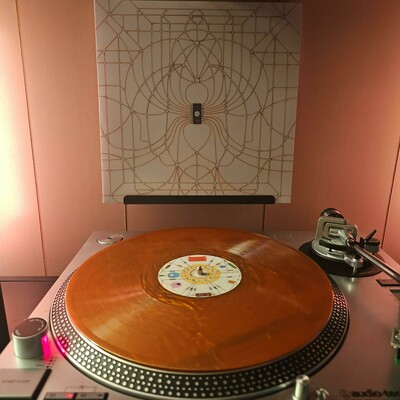 A copper nugget vinyl record sits on a turntable. Behind the turntable, a vinyl album outer sleeve is displayed. The front cover shows a circuit in the middle of a circuit board.