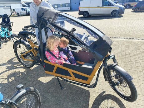 Two children sitting in a cargo bike, while an adult walks behind.