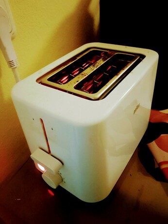 Toast toasting in the toaster, round side down. One can see filaments glowing, and the power light is on. Photographed at a jaunty, artsy farty angle.