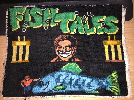 Cross stitching, the top says Fish Tales, in the middle is a man smiling very widely and he has a trophy on each of his sides, below is the same man next to a very large blue fish