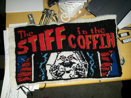 Cross stitching, the top says The Stiff in the Coffin, and below that is a white coffin with a creature with red eyes pushing through it as it comes to life
