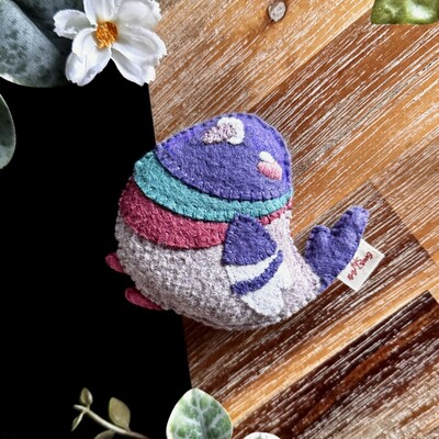 Picture of a handmade felt pigeon plushie photographed on a black and wooden background with a few decorative plants around.
The pigeon is round and looks like it’s sitting down and happily looking up. It’s head, wings and tail are a soft dusky purple, a green stripe and a pink stripe are under its neck and its body is a warm grey color. The tip of its wings are white. A small white squinty eye is embroidered above a pink blush.