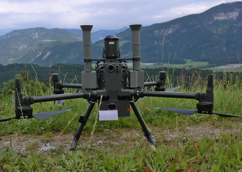 a dji Matrice M300 standing on grassy ground in front of a mountainous background. Under the drone there is a 3d printed rather big black box with radiation shields and a little display, at its sides two 3d printed gray chimnes rise up, these are for particle measurements