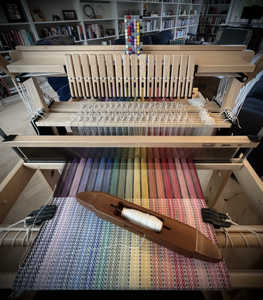 A photo of a Louet Jane 16-shaft handloom warped up with cotton in a rainbow of colours. A handmade wooden boat shuttle sits at an angle on top of the woven fabric