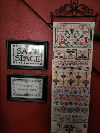 Hanging on a red wall below a staircase:
A long sampler with elaborate counted cross-stitch designs and the words, "OVERTHROW PATRIARCHY."

To the left are 2 framed counted cross-stitch projects:
"SAFE SPACE" in rainbow colors and "Ask me about my feminist agenda" framed in flowers