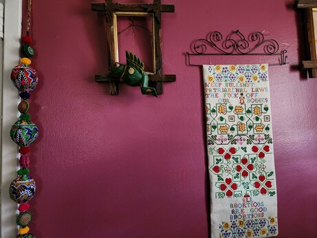 A view of the 2nd sampler and other decor: next to it is an antique picture frame with no picture, just a flying wooden Indonesian frog.

On the other side of the frog: a long string of colorful Indian pompom art.