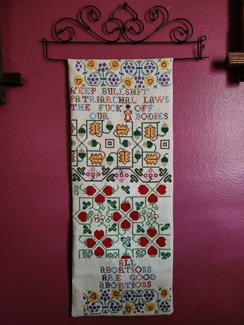 An ornate counted cross-stitch sampler hanging from a curvy black metal hanger

Amidst the designs of flowers, acorns, strawberries, etc. are the words:
"KEEP BULLSHIT PATRIARCHAL LAWS THE FUCK OFF OUR BODIES" and "ALL ABORTIONS ARE GOOD ABORTIONS."