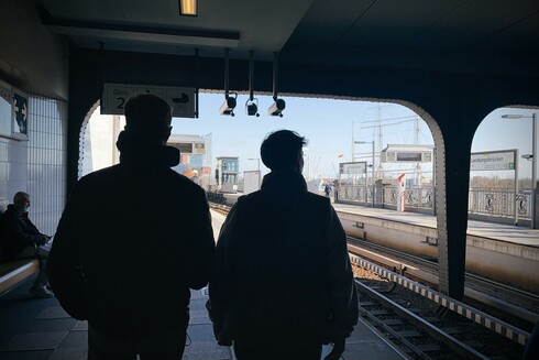 A chest-height photograph along the Landungsbrücken U-Bahn platform in Hamburg. The camera is located under a flat roof made from riveted steel. The area under the roof is dark, but visible. Bright sunshine outside that is "correctly" exposed. The main subject are two male-looking persons walking away from the camera side by side, visible from head to hips. Their silhouettes are framed by the station roof.