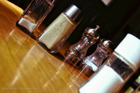 Glass salt and pepper shakers are arranged on a wooden table, casting reflections on the polished surface. Their different shapes and sizes create an interesting composition that highlights their practical design. A lone salt shaker in the foreground looks at the other couples with envy.