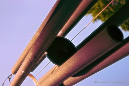 Copper-coloured tubes crisscross a clear blue sky, suspended by strings in an abstract arrangement. A hint of green foliage peeks into the corner, adding a natural contrast to the metallic elements.