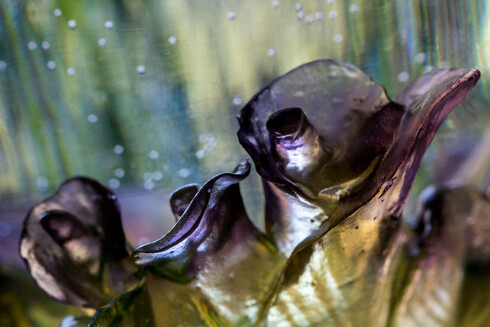 A small detail of glasswork. The photo is split diagonally from bottom left to top right. The bottom half is a row of strange, gold-and-dark-purple, shiny protrusions, resembling alien flowers or creatures. The top half shows faint, light green diagonal stripes—perhaps a forest or grass far away?