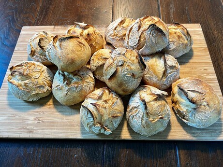 Auf einem dunklen Holztisch liegt ein Holzbrett. Darauf liegen zahlreiche frisch gebackene Brötchen mit rustikalem Ausbund