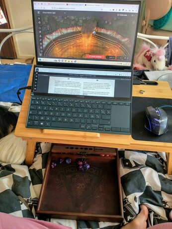 A laptop propped on a bed table with a map on one screen and a second screen above the keyboard has a PDF of text on it. Underneath the mini table is a wooden dice tray with a set of purple dice in it.