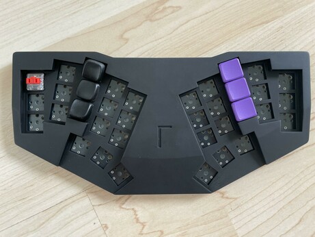 Overhead shot of a monoblock split keyboard (the Gamma Omega case design) in black resin. One “red pro” low profile choc v1 switches top left, the right middle finger column in black are 3D printed resin sculpted KLP Lamé keycaps (rising above the case), the right middle finger column in purple are MBK “Violet” keycaps (flush with the case)