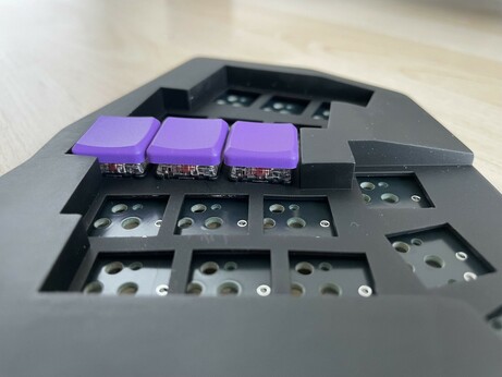 Side view of a monoblock split keyboard (the Gamma Omega case design) in black resin. One column fitted with choc v1 switches and MBK “Violet” keycaps in purple (flush with the case)