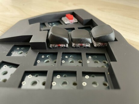 Side view of a monoblock split keyboard (the Gamma Omega case design) in black resin. One red low profile choc v1 switches top corner, and the right middle finger column alone are filled. This has red switches with black 3D printed resin sculpted KLP Lamé keycaps. The photo angle shows how much the top and bottom keys extend upwards from the case surface to create a well for the finger.