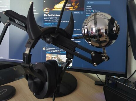 Photo of Leraje's headphones (complete with fake Devil horns) and microphone against a backdrop of a PC monitor showing The Devil's Library Bluesky profile page.