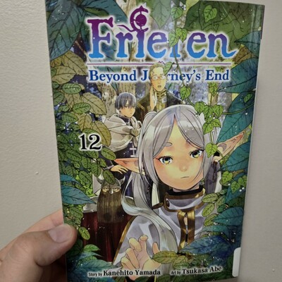 Photograph of a white person's hand holding Frieren: Beyond Journey's End, volume 12. Image shows the titular Frieren centralized peeking through some branches with a party of adventurers visible behind her.