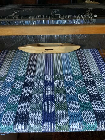 Photo of weaving in process on the loom. It is stripes of various blues and green with white polka dots.