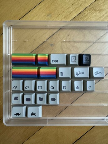 A keycap tray with icon mod keycaps in it.
There's a few with the Apple logo colours, one that says "1984", happy and sad Mac ones. From MacPaint: eraser, lasso, rectangle marquee, paint bucket, hand, paint brush, pencil, the shape tools, the "turtle" and "rabbit" icons from the control panel for mouse speed, a trash can, and two others representing documents (but not from the original Mac system as far as I can tell).