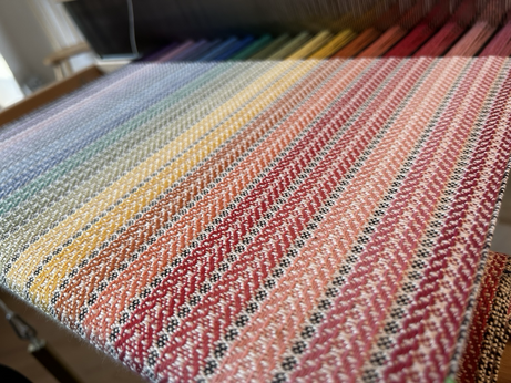 An oblique photo of spiral twill fabric in a rainbow of colours, in progress on a hand loom