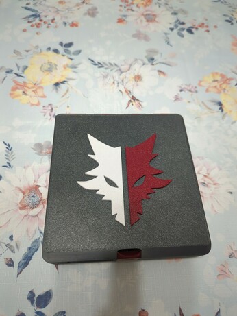 A Gridfinity box, whose outer shell is printed from a dark grey plastic. The box is currently closed, with only the top face visible, which is textured like a print off a textured PEI sheet would be. On the top face is a logo shaped like a wolf, in two halves: one is white, the other is a dark red. The latch is also visible, and it's in the same dark red as half of the wolf.