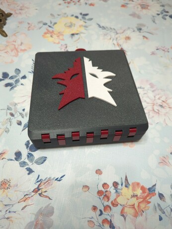 A Gridfinity box, whose outer shell is printed from a dark grey plastic, viewed from the back. The box is currently closed, with only the top face visible, which is textured like a print off a textured PEI sheet would be. On the top face is a logo shaped like a wolf, in two halves: one is white, the other is a dark red. The hinges are also visible, and are printed in the same dark red as half of the wolf.