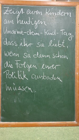 Auf grüner Tafel steht mit weißer Kreide:
Zeigt euren Kindern am heutigen Umarme-dein-Kind-Tag, dass ihr sie liebt, wenn sie denn schon die Folgen eurer Politik ausbaden müssen.