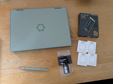 A picture of the Framework 12 laptop closed on a table.  It's in sage green with a dark green cog logo.  Next to it is a sage green screwdriver, 16GB RAM, four USB-C modules, and an SSD.