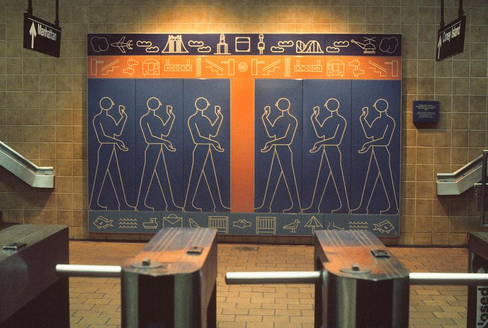Photograph of the artwork 'Kings Highway Hieroglyphs' (1987) by Rhoda Yohai Andors at the Kings Hwy subway station. The mural features six outlined human figures standing in profile, each with one arm raised. The figures are each set on their own vertical panel with a blue background, and the three on the left face the three on the right, separated by an orange stripe. Above and below the figures are rows of stylized pictograms of things like clouds, an airplane, a bridge, a roller coaster, a helicopter, fish, and birds, modern visual symbols echoing ancient hieroglyphs. A plaque with more information about the artwork is visible but illegible. Subway turnstiles are visible in the foreground, with directional signs for 'Manhattan' and 'Coney Island' hanging above. Photo by Rhoda Yohai Andors via MTA Arts & Design at https://www.mta.info/agency/arts-design/collection/Kings-Highway-Hieroglyphs.