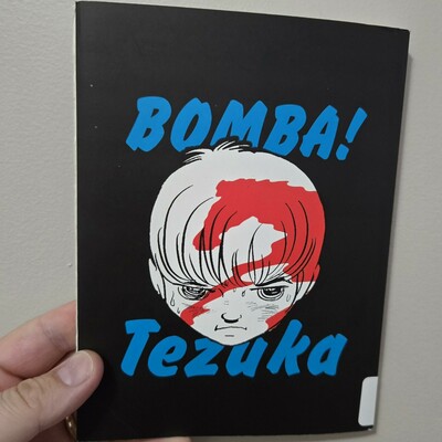 Photograph of a white person's hand holding a book against a light colored wall. The book has a black cover with the words "Bomba!" and "Tezuka" in blue, text above and below a black and white teenage boy's face with a red silhouette of a horse shown within it.