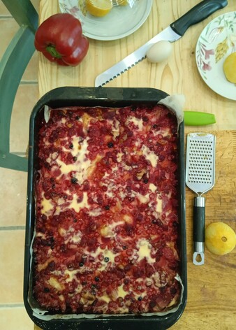A large tray bake with cheesecake and red berry topping, resting on a table to cool. A lemon grater and half lemon sit on the breadboard to the right, various plates and knives above.