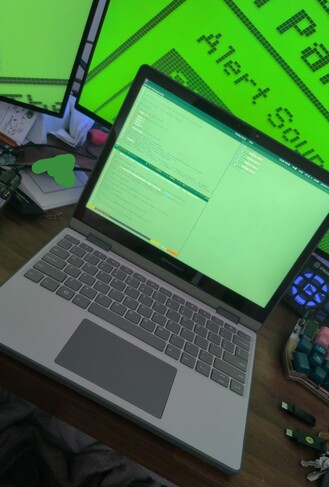 A picture of my green framework 12 laptop open on my desk.  It has a grey keyboard cover.  The screen is displaying my new Sway environment with a dark green top bar and two terminal windows open side by side, both styled with a light green background.  In the backdrop my desktop monitor displays a green screensaver.  My desk has assorted niknaks around, including a Yubikeys and a Stream Deck.  I've scribbled over an open notebook with green using the pen tool.