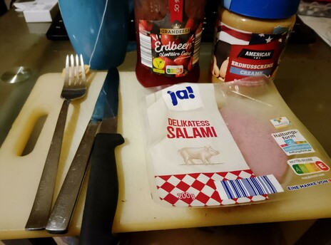 Ingredients for Sweet & Sour Pork Sandwiches:
Strawberry preserves, creamy peanut butter, and salami sit on an acrylic cutting board, along with cutlery. A fresh blue cuppa steeps alongside.