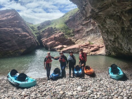 4 happy kayakers & 5 kayaks on pebbly beach in grotto in cliffs.
