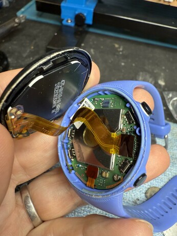 An open smartwatch is held in a hand, showing its internal components, including a green circuit board and various connections. The watch has a blue rubber strap.